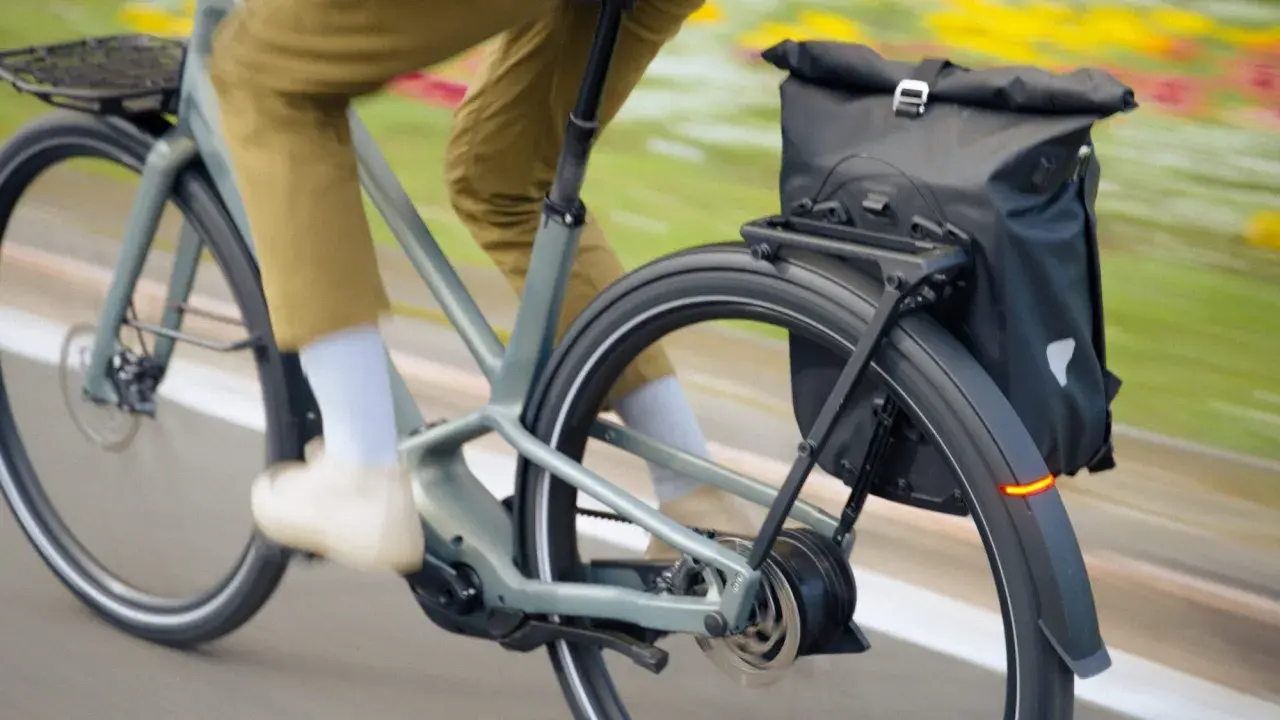 Un cycliste en pantalon kaki roule sur un vélo électrique gris, sacoche noire à l'arrière. Facile à utiliser, ce vélo offre une assistance douce.