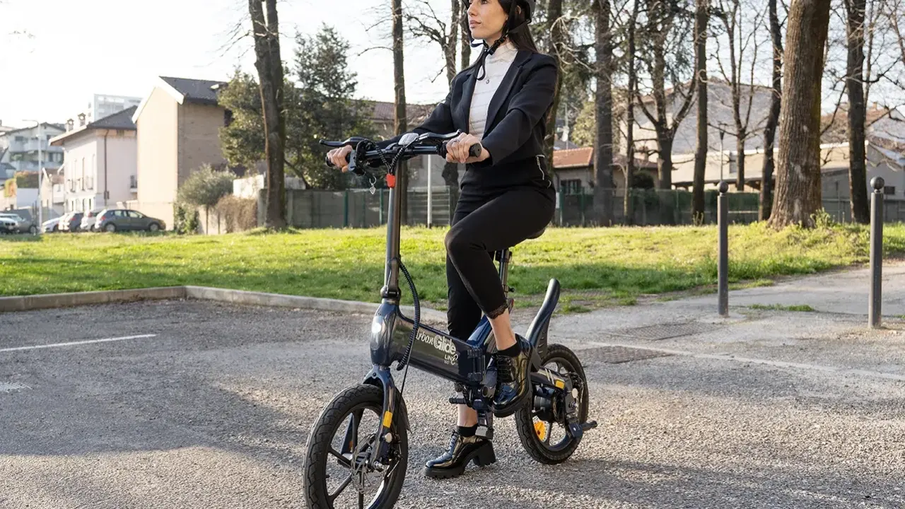 Femme en costume, casque, assise sur une trottinette Urban Glide qui n'avance plus. Elle semble dépitée.