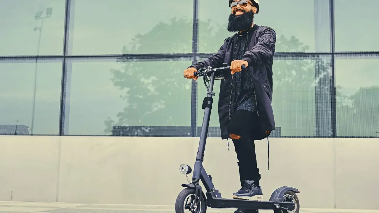 Un homme barbu en casque et lunettes roule sur une trottinette électrique devant un mur de verre. La question "peut-on rouler sur la route avec une trottinette électrique" est pertinente.