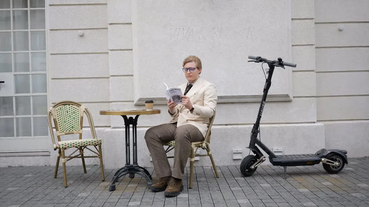 Un homme lit un livre à côté d'une trottinette électrique, prêt à reprendre sa route à toute vitesse.