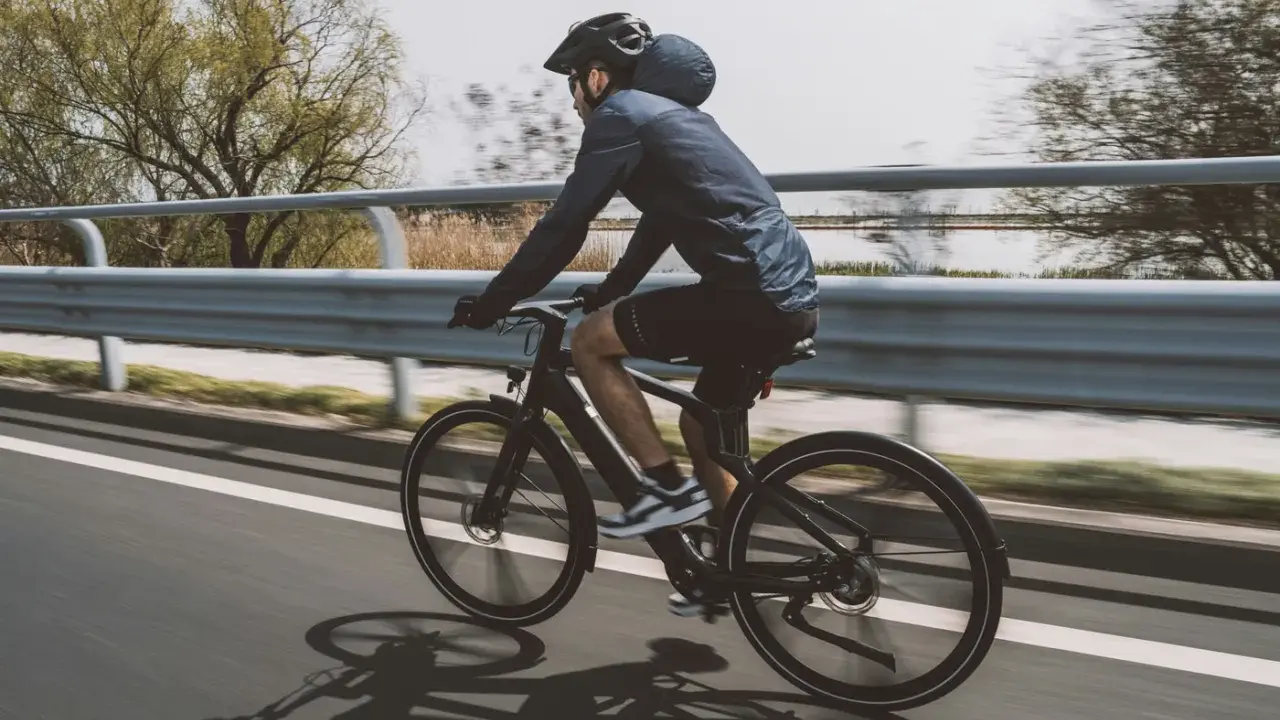 Un cycliste sur un vélo électrique qui se recharge en roulant, longeant une voie rapide bordée d'eau et d'arbres.