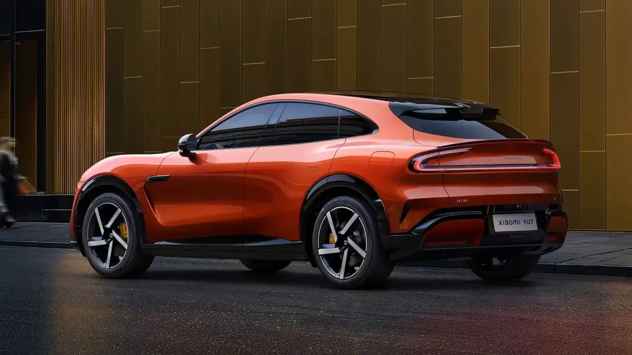 La **Xiaomi car** orange vif, un SUV coupé futuriste, se gare devant un bâtiment moderne aux reflets dorés.