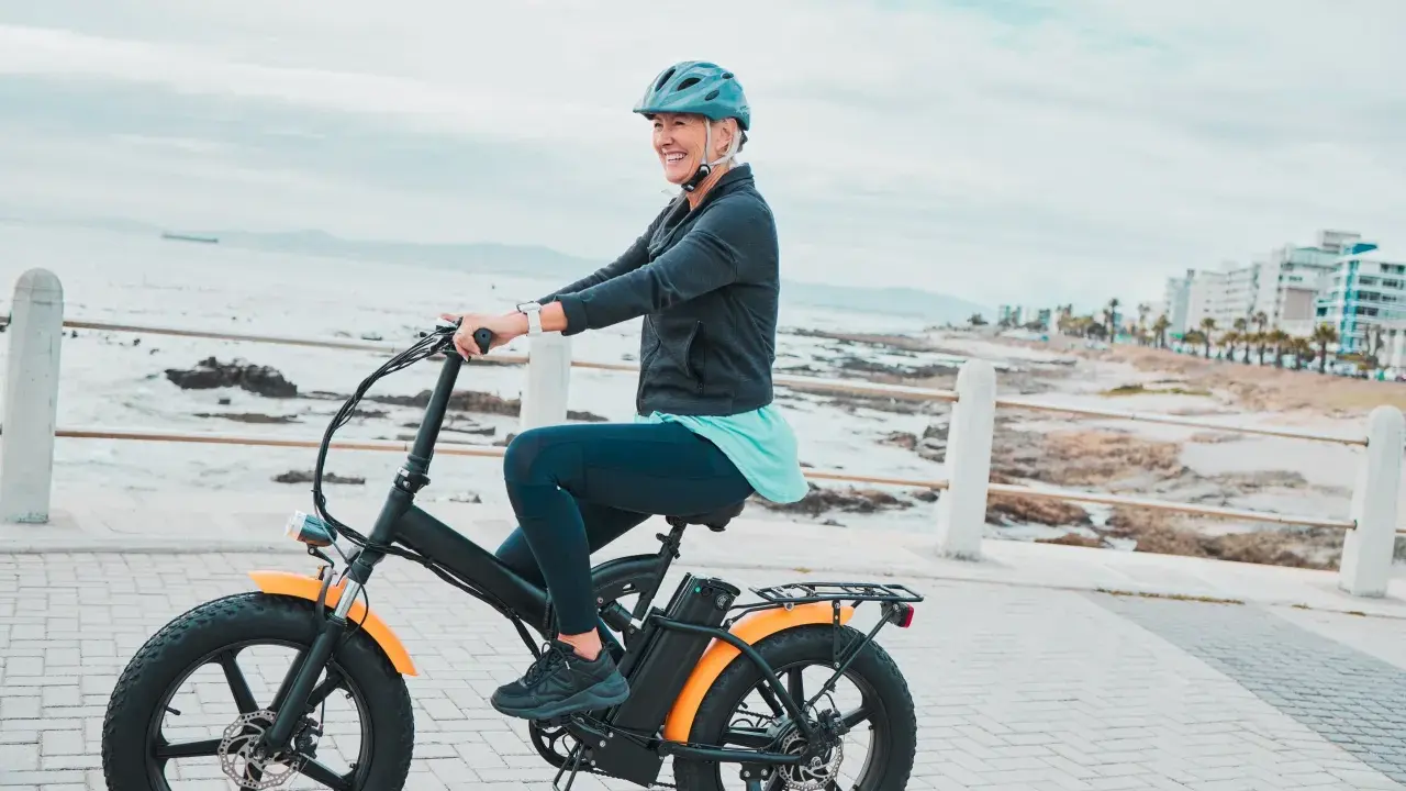 Une femme souriante roule sur un vélo électrique, profitant de la vue sur la mer. Le poid du vélo électrique ne l'empêche pas de profiter de sa balade.