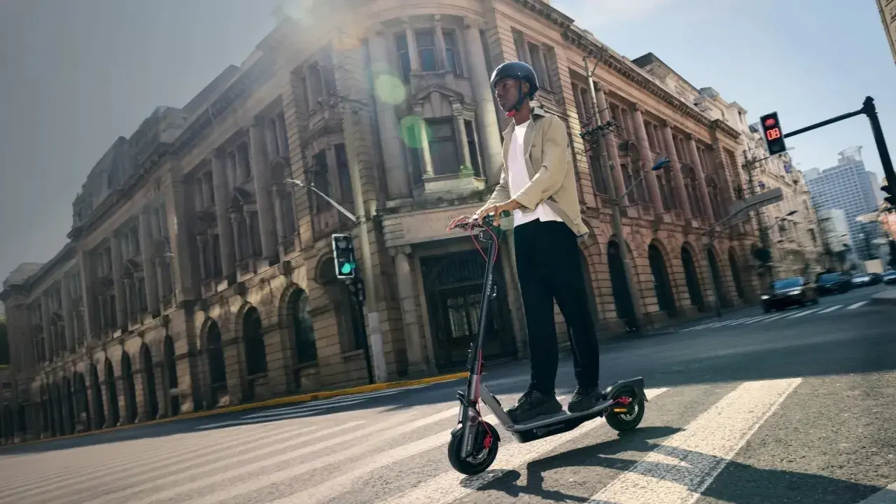 Un homme en casque roule sur la trottinette électrique la plus rapide du monde, traversant une rue bordée de bâtiments historiques.