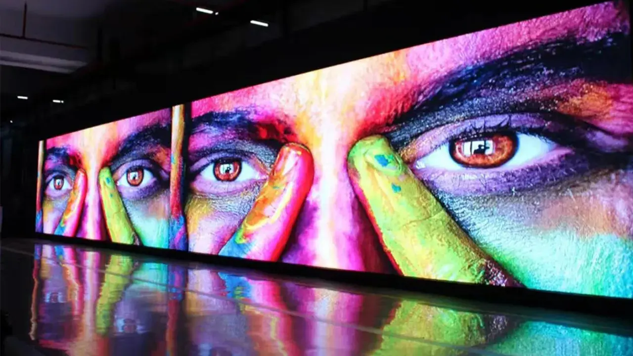 Un écran géant affiche un visage aux yeux intenses, couvert de peinture colorée. La dalle IPS ou LED rend les couleurs vibrantes et le reflet sur le sol ajoute à l'effet visuel.