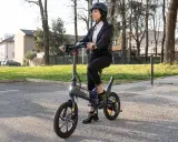 Femme en costume, casque, assise sur une trottinette Urban Glide qui n'avance plus. Elle semble dépitée.