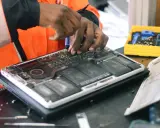 Réparation d'un ordinateur portable ouvert, mains expertes utilisant un tournevis. Une boîte à outils jaune est visible, prête pour la prochaine étape, sans deee taxe.