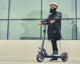 Un homme barbu en casque et lunettes roule sur une trottinette électrique devant un mur de verre. La question "peut-on rouler sur la route avec une trottinette électrique" est pertinente.