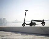 Une trottinette électrique s'allume mais ne bouge pas, posée sur un muret face à une ville et un pont.