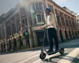 Un homme en casque roule sur la trottinette électrique la plus rapide du monde, traversant une rue bordée de bâtiments historiques.