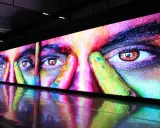 Un écran géant affiche un visage aux yeux intenses, couvert de peinture colorée. La dalle IPS ou LED rend les couleurs vibrantes et le reflet sur le sol ajoute à l'effet visuel.