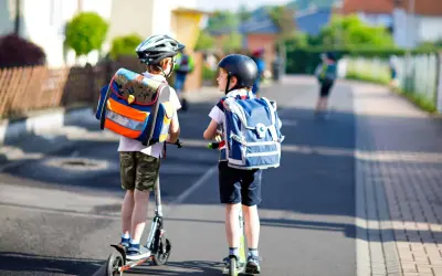 Deux enfants casqués, sacs à dos sur les épaules, se déplacent en trottinette. L'âge trottinette électrique est idéal pour ces jeunes écoliers.