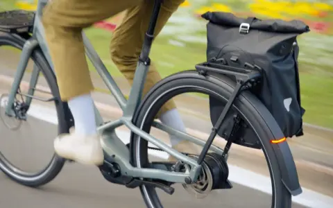 Un cycliste en pantalon kaki roule sur un vélo électrique gris, sacoche noire à l'arrière. Facile à utiliser, ce vélo offre une assistance douce.