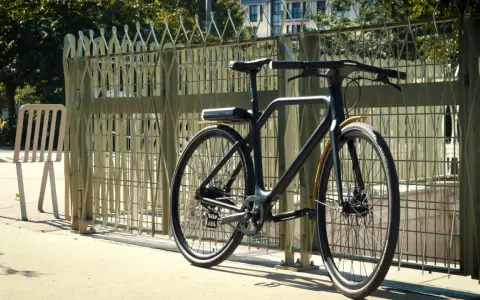 Un vélo angell noir, au design épuré, est garé près d'une grille métallique. Une chaise blanche est à gauche.