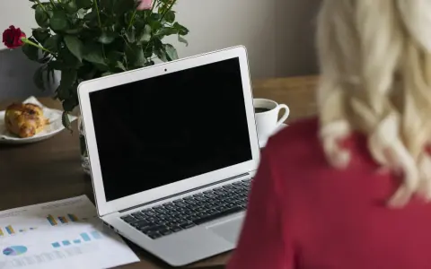 Un ordinateur portable ouvert sur un bureau en bois, avec une tasse de café, des roses et un croissant. Le branchement PC est prêt pour le travail.