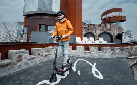 Un homme en casque et veste orange teste la vitesse de sa trottinette électrique devant un bâtiment de style médiéval moderne.