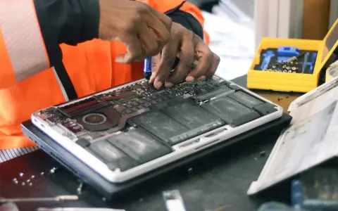 Réparation d'un ordinateur portable ouvert, mains expertes utilisant un tournevis. Une boîte à outils jaune est visible, prête pour la prochaine étape, sans deee taxe.