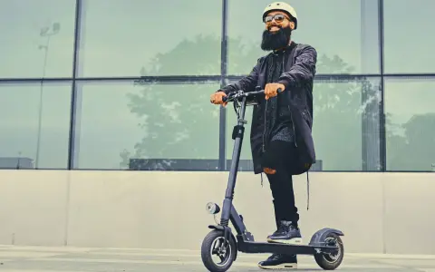 Un homme barbu en casque et lunettes roule sur une trottinette électrique devant un mur de verre. La question "peut-on rouler sur la route avec une trottinette électrique" est pertinente.