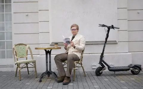 Un homme lit un livre à côté d'une trottinette électrique, prêt à reprendre sa route à toute vitesse.