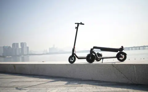 Une trottinette électrique s'allume mais ne bouge pas, posée sur un muret face à une ville et un pont.