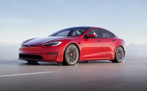 Une Tesla Model S Plaid rouge vif file sur une route côtière, le soleil se reflétant sur sa carrosserie élégante.