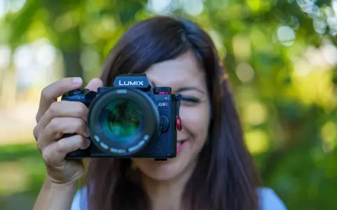 Une femme souriante tient un appareil photo Lumix G9II devant son visage. Un guide pour comment choisir son appareil photo.