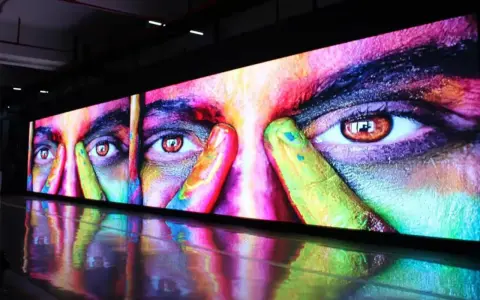 Un écran géant affiche un visage aux yeux intenses, couvert de peinture colorée. La dalle IPS ou LED rend les couleurs vibrantes et le reflet sur le sol ajoute à l'effet visuel.