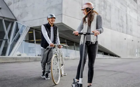 Un homme à vélo et une femme en trottinette, tous deux portant un casque obligatoire, discutent devant un bâtiment moderne.
