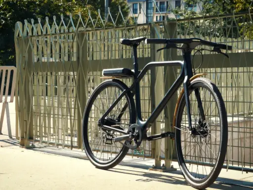 Un vélo angell noir, au design épuré, est garé près d'une grille métallique. Une chaise blanche est à gauche.