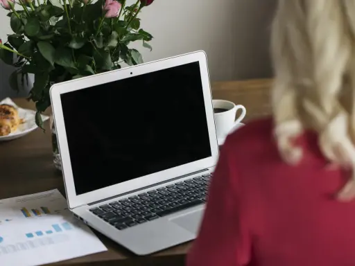 Un ordinateur portable ouvert sur un bureau en bois, avec une tasse de café, des roses et un croissant. Le branchement PC est prêt pour le travail.