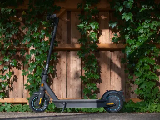 Trottinette électrique noire, prête à l'aventure, avec une puissance impressionnante, posée devant un mur de bois couvert de végétation.