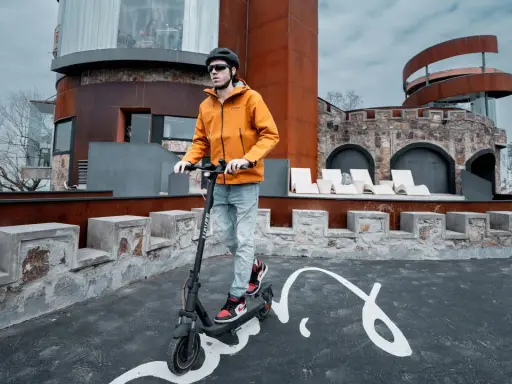 Un homme en casque et veste orange teste la vitesse de sa trottinette électrique devant un bâtiment de style médiéval moderne.