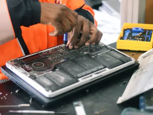 Réparation d'un ordinateur portable ouvert, mains expertes utilisant un tournevis. Une boîte à outils jaune est visible, prête pour la prochaine étape, sans deee taxe.