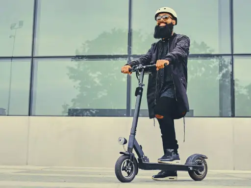 Un homme barbu en casque et lunettes roule sur une trottinette électrique devant un mur de verre. La question "peut-on rouler sur la route avec une trottinette électrique" est pertinente.