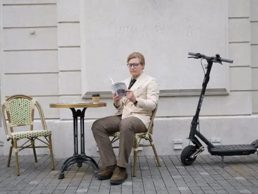 Un homme lit un livre à côté d'une trottinette électrique, prêt à reprendre sa route à toute vitesse.