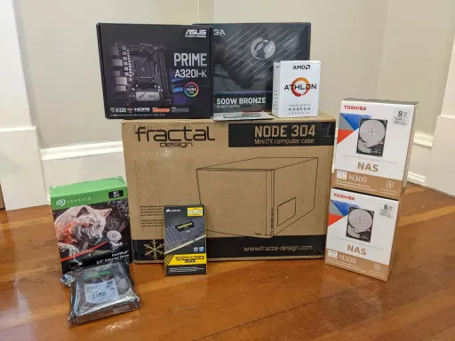 Composants pour installer un NAS : carte mère ASUS, processeur AMD Athlon, boîtier Fractal Design, alimentation, RAM Corsair, et deux disques durs Toshiba 8 To.