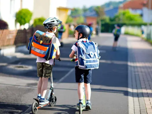 Deux enfants casqués, sacs à dos sur les épaules, se déplacent en trottinette. L'âge trottinette électrique est idéal pour ces jeunes écoliers.