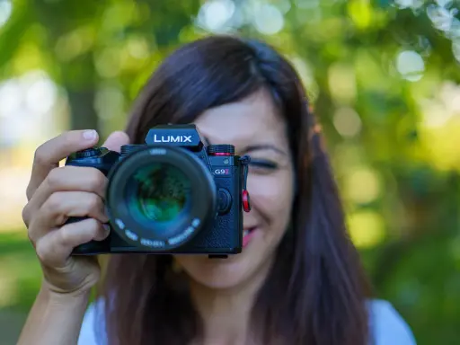 Une femme souriante tient un appareil photo Lumix G9II devant son visage. Un guide pour comment choisir son appareil photo.