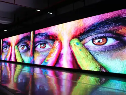 Un écran géant affiche un visage aux yeux intenses, couvert de peinture colorée. La dalle IPS ou LED rend les couleurs vibrantes et le reflet sur le sol ajoute à l'effet visuel.