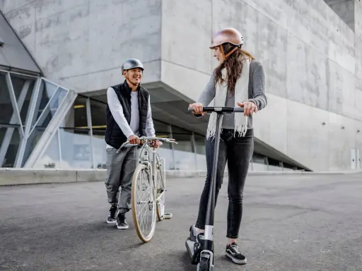 Un homme à vélo et une femme en trottinette, tous deux portant un casque obligatoire, discutent devant un bâtiment moderne.