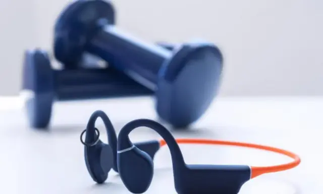 Un casque à conduction osseuse bleu et orange, prêt pour votre prochaine séance de sport, avec des haltères bleus en arrière-plan.