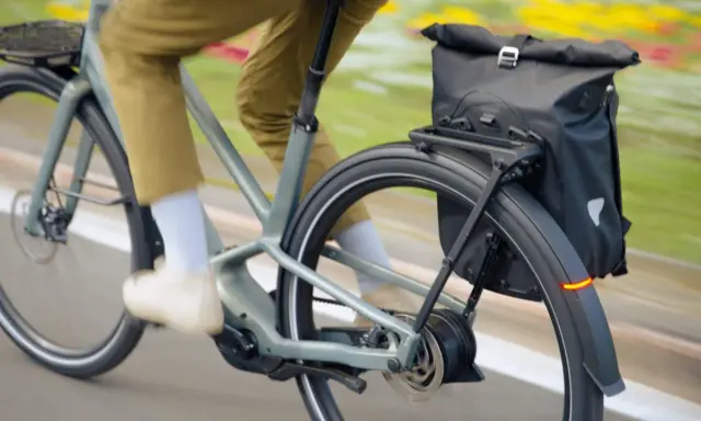 Un cycliste en pantalon kaki roule sur un vélo électrique gris, sacoche noire à l'arrière. Facile à utiliser, ce vélo offre une assistance douce.