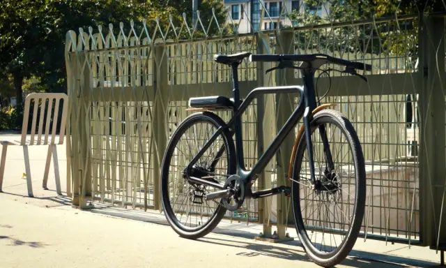 Un vélo angell noir, au design épuré, est garé près d'une grille métallique. Une chaise blanche est à gauche.