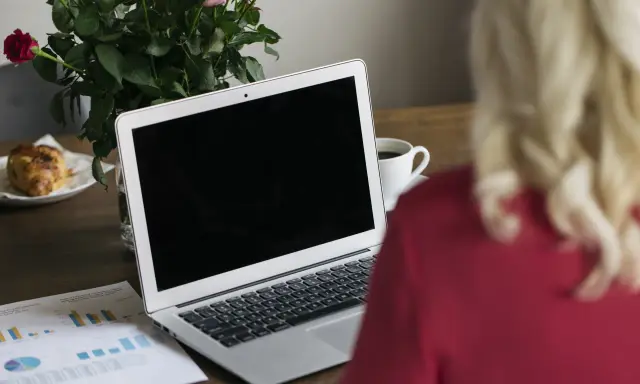 Un ordinateur portable ouvert sur un bureau en bois, avec une tasse de café, des roses et un croissant. Le branchement PC est prêt pour le travail.