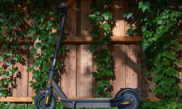 Trottinette électrique noire, prête à l'aventure, avec une puissance impressionnante, posée devant un mur de bois couvert de végétation.