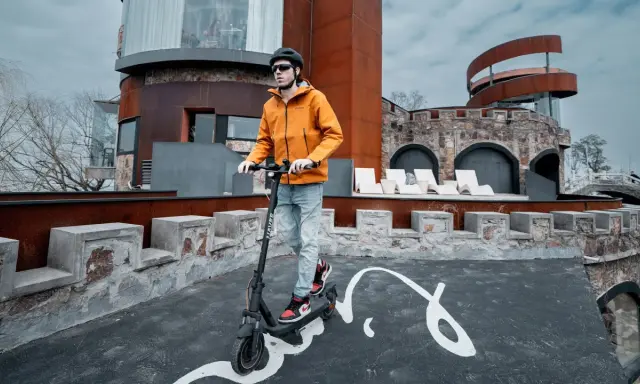 Un homme en casque et veste orange teste la vitesse de sa trottinette électrique devant un bâtiment de style médiéval moderne.
