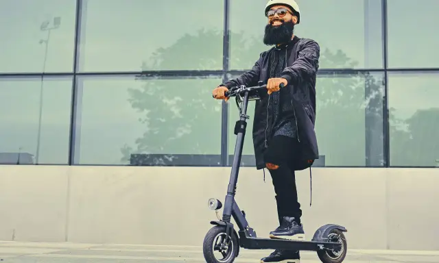 Un homme barbu en casque et lunettes roule sur une trottinette électrique devant un mur de verre. La question "peut-on rouler sur la route avec une trottinette électrique" est pertinente.