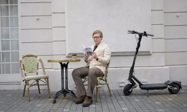 Un homme lit un livre à côté d'une trottinette électrique, prêt à reprendre sa route à toute vitesse.