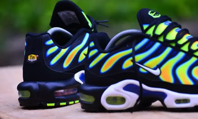Paire de baskets Nike Air Max Plus TN, noires avec des vagues dégradées de jaune, orange et bleu.