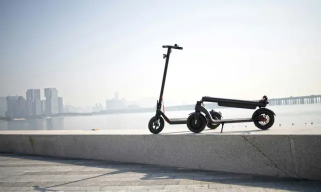 Une trottinette électrique s'allume mais ne bouge pas, posée sur un muret face à une ville et un pont.